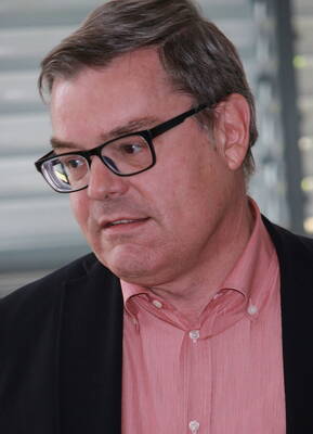 Mittelalterlicher Mann mit Brille und grauem Haar im Profil, in rosa Hemd und dunklem Sakko, schaut nachdenklich zur Seite.