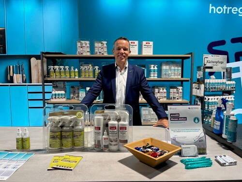 Messeaussteller im Anzug präsentiert an einem Stand der Marke hotrega verschiedene Reinigungs- und Pflegeprodukte, Infomaterial und Werbegeschenke vor einem blau gestalteten Hintergrund.