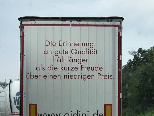 Rückseite eines Lkw mit dem roten Werbespruch „Die Erinnerung an gute Qualität hält länger als die kurze Freude über einen niedrigen Preis“ vor bewölktem Himmel.