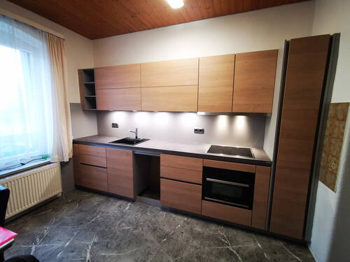 Modern eingerichtete Einbauküche mit holzfarbenen Fronten, grauer Arbeitsplatte, Induktionskochfeld, Backofen, Spülbecken und Unterbau-LED-Beleuchtung entlang der geraden Küchenzeile.
