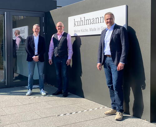Drei lächelnde Männer in Business‑Casual-Kleidung stehen im Sonnenschein vor dem Firmeneingang von Kuhlmann Küchen neben einem großen Firmenlogo an der dunklen Außenwand.