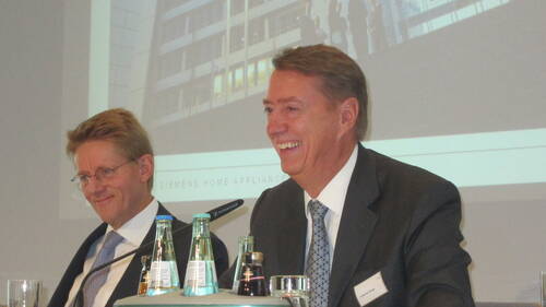 Zwei lächelnde Geschäftsleute im Anzug sitzen mit Mikrofonen und Getränkeflaschen vor sich auf einem Podium während einer Präsentation in einem Konferenzraum.