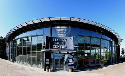 Modernes, halbrundes Glasgebäude des „Italian Yacht Centre“ mit großem Eingangsportal und spiegelnden Fassaden unter klarem blauem Himmel