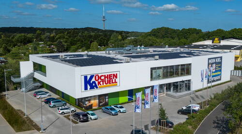 Modernes Küchenstudio-Gebäude mit großem Küchen Aktuell Logo, Solaranlage auf dem Dach und parkenden Autos vor grüner Waldkulisse.