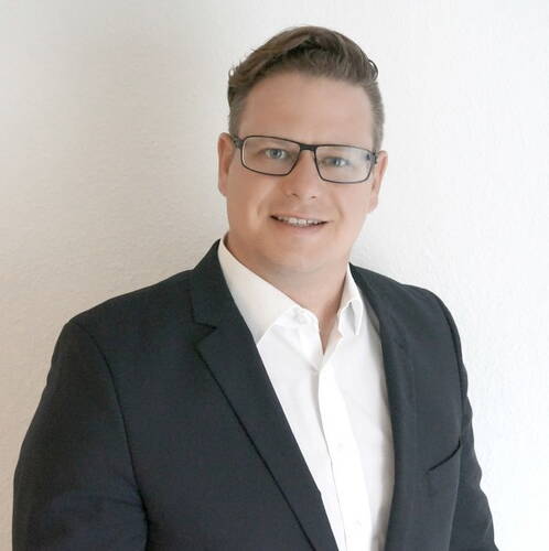 Geschäftsmann mit Brille im dunklen Anzug und weißem Hemd lächelt vor einer hellen Wand in die Kamera