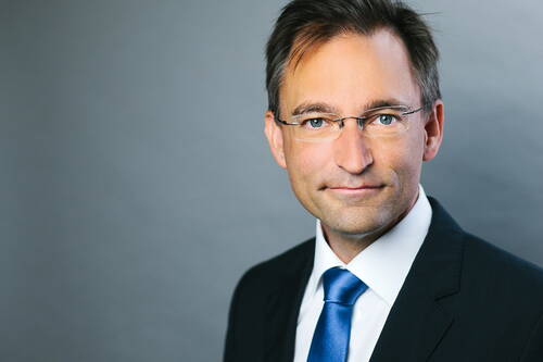 Seriöser Geschäftsmann mit Brille im Anzug und blauer Krawatte blickt lächelnd in die Kamera vor neutral grauem Hintergrund