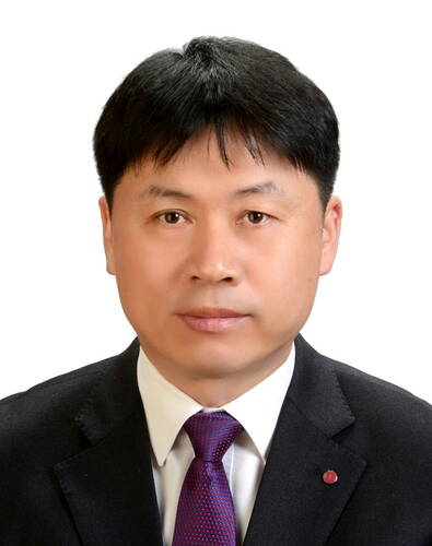 Mittelalterlicher asiatischer Mann im Anzug mit weißem Hemd und violetter Krawatte blickt mit neutralem Gesichtsausdruck direkt in die Kamera vor weißem Hintergrund.