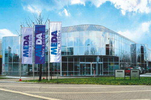 Modernes, rundes Küchenfachgeschäft mit gläserner Fassade und hohen Fahnenmasten mit MEDA-Logo vor blauem Himmel