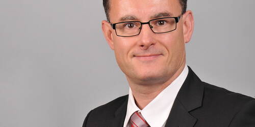 Geschäftsmann mit Brille im dunklen Anzug und gestreifter Krawatte lächelt vor neutral grauem Hintergrund.