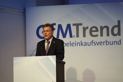 Mittelälterer Mann im Anzug hält eine Rede am Rednerpult vor einer Präsentationswand mit dem Logo „GfMTrend Möbeleinkaufsverbund“.