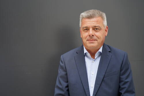 Mittelalterlicher Mann im blauen Anzug mit gestreiftem Hemd steht vor einer einfarbig grauen Wand und blickt ruhig in die Kamera.