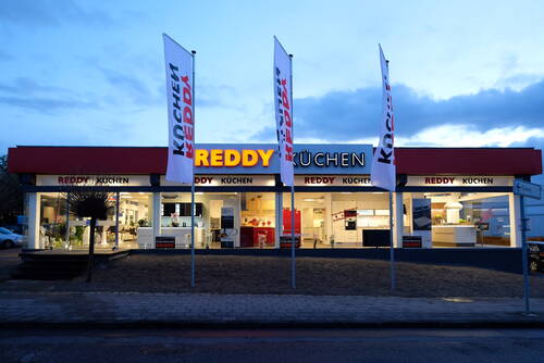 Abendliche Außenansicht eines hell erleuchteten Reddy-Küchenstudios mit großen Schaufenstern und hohen Werbefahnen entlang des Gehwegs