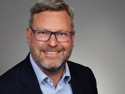 Mittelalterlicher Mann mit grauem Haar, Bart und blauer Brille lächelt im Business-Porträt vor neutral grauem Hintergrund