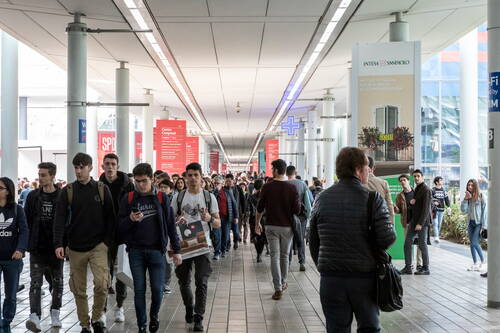 Menschenmenge junger und älterer Besucher geht durch einen überdachten Messegang mit zahlreichen roten Hinweisschildern und Werbetafeln an den Seiten.