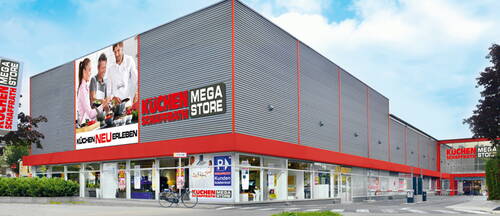 Moderner Küchenschaupark-Megastore an einer Straßenecke mit großer Glasfront, auffälliger roter Umrandung und Werbebanner mit kochender Familie an der Fassade