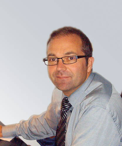 Mittelalterlicher Mann mit Brille und Krawatte sitzt seitlich an einem Schreibtisch und blickt freundlich in die Kamera