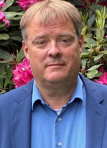 Mittelalterlicher Mann im blauen Sakko und Hemd steht vor blühenden pinken Rhododendronsträuchern und blickt ruhig in die Kamera.