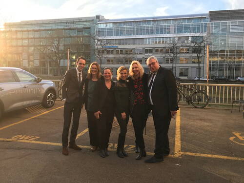 Sechs elegant gekleidete Personen posieren lächelnd im Abendlicht auf einem Parkplatz vor einem modernen Bürogebäude.