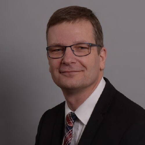 Mittelälterer Mann mit Brille im Anzug und kariertem Schlips lächelt vor neutral grauem Hintergrund