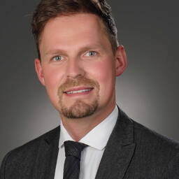 Junger Mann mit kurzem braunem Haar und Bart, der im grauen Anzug mit Krawatte vor neutralem dunklem Hintergrund freundlich in die Kamera lächelt.