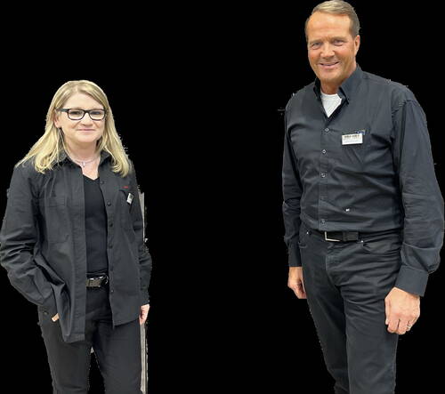 Zwei Mitarbeitende in schwarzer Arbeitskleidung lächeln in die Kamera und tragen Namensschilder vor schwarzem Hintergrund.