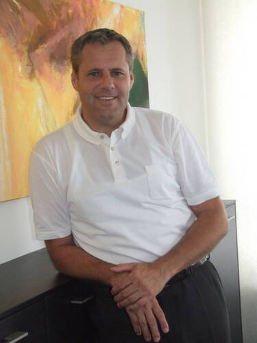 Mittelälterlicher Mann in weißem Poloshirt lehnt lächelnd an einem dunklen Sideboard vor einem bunten abstrakten Gemälde in einem hellen Raum