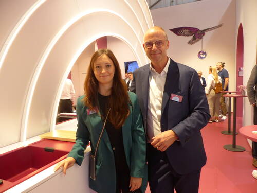 Zwei Messebesucher in Businesskleidung posieren lächelnd in einem modern gestalteten, pink beleuchteten Ausstellungsstand mit geschwungenen Lichtbögen im Hintergrund.