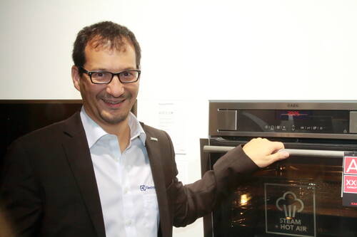 Mann im Anzug lächelt und präsentiert einen modernen AEG-Einbaubackofen mit Dampffunktion im Ausstellungsraum
