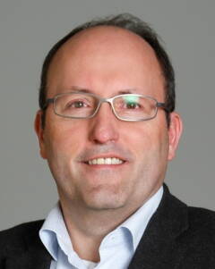 Mittelalterlicher Mann mit Brille und kurzem Haar lächelt in die Kamera und trägt Hemd und dunkles Sakko vor neutral grauem Hintergrund