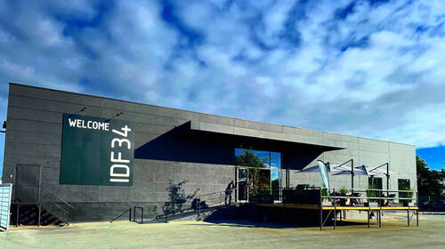 Moderner, grauer Flachbau mit großer „WELCOME IDF34“-Beschriftung, verglastem Eingangsbereich und Terrasse mit Sonnenschirmen unter leicht bewölktem Himmel