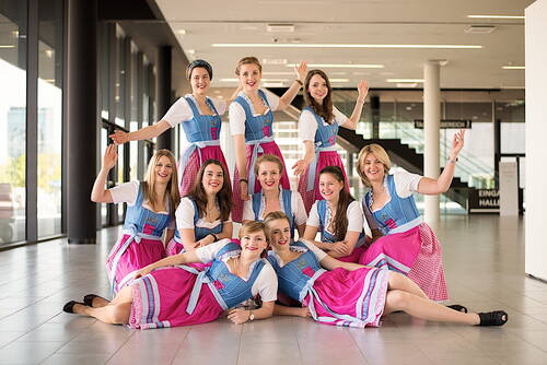 Neun junge Frauen in traditionellen Dirndln mit blauen Miedern und pinken Schürzen posieren fröhlich in einem modernen, lichtdurchfluteten Foyer und strecken lachend die Arme in die Höhe.