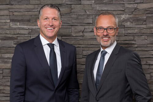 Zwei lächelnde Geschäftsleute im Anzug stehen vor einer grauen Steinwand und blicken direkt in die Kamera.