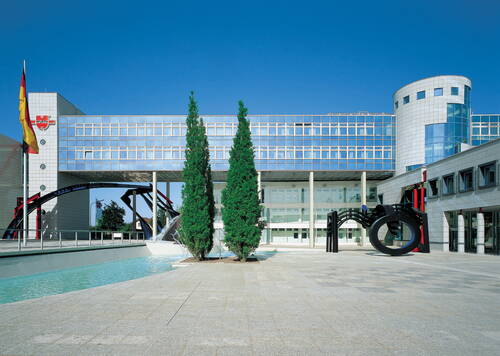 Moderner Bürokomplex mit verglaster Fassade, Wasserbecken, zwei hohen Nadelbäumen und schwarzer Metallskulptur im weitläufigen Innenhof unter blauem Himmel