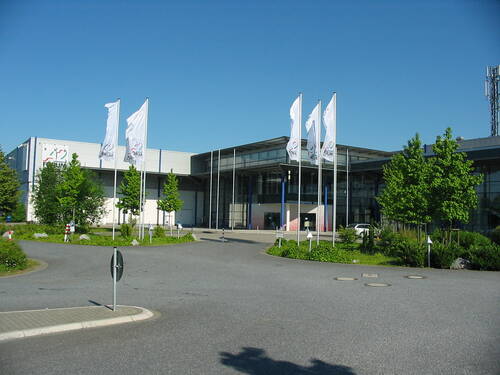 Modernes Messe- und Veranstaltungsgebäude mit Glasfassade, umgeben von Grünflächen und mehreren hohen weißen Fahnen an einem sonnigen Tag