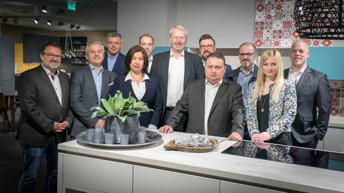 Gruppe von zwölf Geschäftsleuten steht in einer modernen Ausstellungsküche hinter einer hellen Kücheninsel mit Dekotablett und grüner Pflanze