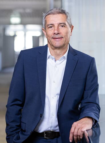 Älterer Mann im blauen Anzug lehnt entspannt an einem Geländer in einem modernen, hellen Büroflur und blickt freundlich in die Kamera