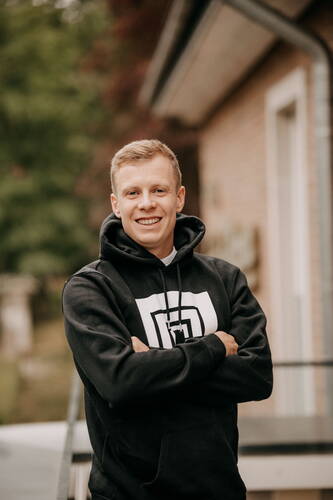 Junger Mann in schwarzem Hoodie mit grafischem Logo steht lächelnd mit verschränkten Armen im Freien vor einem Gebäude