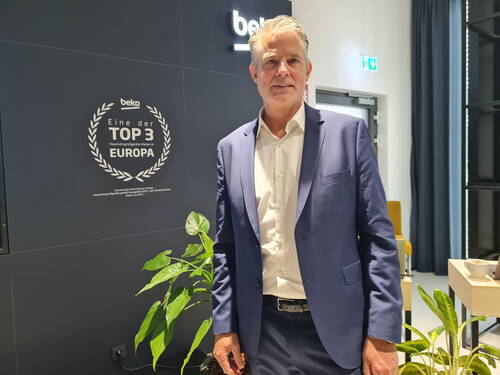 Geschäftsmann im blauen Anzug steht in modernem Showroom vor einer schwarzen Wand mit „Eine der Top 3 Haushaltsgroßgeräte-Marken in Europa“-Auszeichnung und grünen Zimmerpflanzen.
