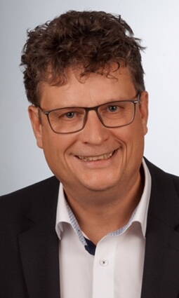 Mittelalterlicher Mann mit lockigem braunem Haar, Brille und freundlichem Lächeln in dunklem Anzug und weißem Hemd vor neutral grauem Hintergrund