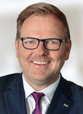 Lächelnder Mann mittleren Alters mit Brille im blauen Anzug und lila Krawatte vor hellem Hintergrund