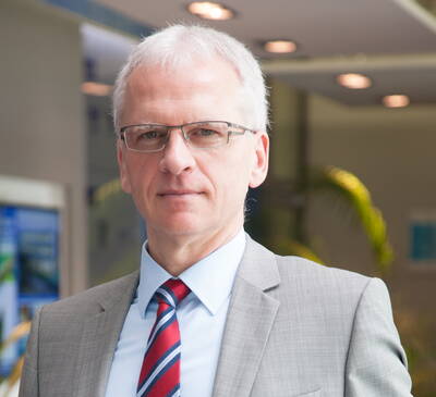 Älterer Geschäftsmann mit grauen Haaren, Brille und grauem Anzug mit rot-blau gestreifter Krawatte steht in einem modernen Bürogebäude und blickt direkt in die Kamera.