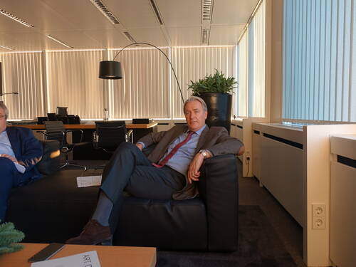 Älterer Mann im Anzug sitzt entspannt auf einem schwarzen Ledersofa in einem modernen Büro mit Jalousien und Konferenztisch im Hintergrund