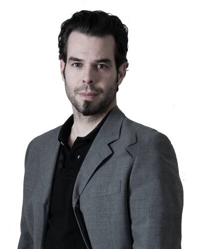 Mann mit dunklem Haar und markantem Kinnbart trägt grauen Blazer über schwarzem Polohemd und blickt ernst in die Kamera vor weißem Hintergrund.