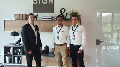 Drei lächelnde Geschäftsleute in weißem Hemd und Anzug stehen in einem modern eingerichteten Büro vor einem Holzregal mit Dekoobjekten und dem Schriftzug „SIGN“ an der Wand