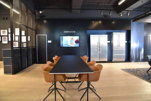 Moderner Showroom mit langem Konferenztisch, ledernen Drehstühlen und wandintegrierten Edelstahl-Kühlschränken vor dunkelblauer Rückwand