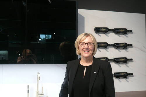 Lächelnde Frau mit Brille in schwarzem Blazer steht in moderner Küche vor einer Wand mit horizontal gelagerten Weinflaschen
