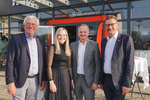 Vier lächelnde Personen in Businesskleidung posieren bei Sonnenschein vor einem Eingang mit dem Schriftzug „Herzlich Willkommen“ bei einer Veranstaltung.