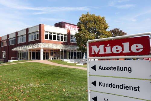 Miele Firmengebäude mit begrüntem Vorplatz und Weg, im Vordergrund ein Hinweisschild zu Ausstellung und Kundendienst