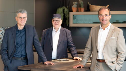 Drei lächelnde Männer in Anzügen stehen locker an einer Kücheninsel in einem modernen, stilvoll eingerichteten Showroom.