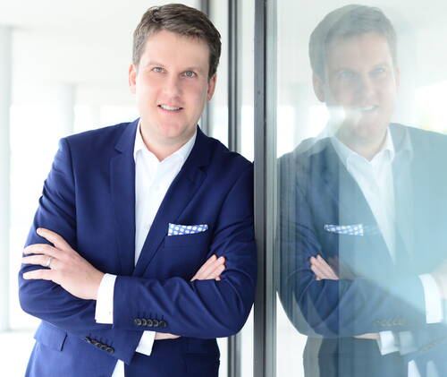 Mann im blauen Business-Anzug steht lächelnd mit verschränkten Armen an einer Glaswand, in der sich sein Oberkörper deutlich spiegelt.